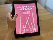 Eine Hand hält ein Tablet. Das Tablet zeigt das Bildungsmaterial "Verschwörungserzählungen. Methoden zum Umgang im Unterricht und in der außerschulischen Bildungsarbeit".