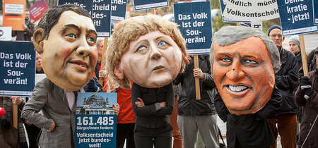 Darsteller mit Großmasken von Gabriel, Seehofer und Merkel