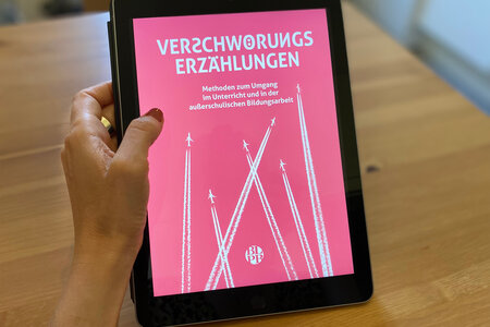 Verschwörungserzählungen. Das Buchcover ist im Tablet zu sehen. 