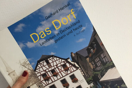 Eine Hand hält das Buch "Das Dorf" vor eine weiße Wand"