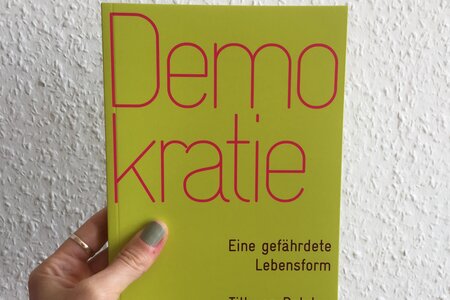Eine Hand hält das Buch "Demokratie. Eine gefährdete Lebensform" von Till van Rahden vor eine weiße Wand.