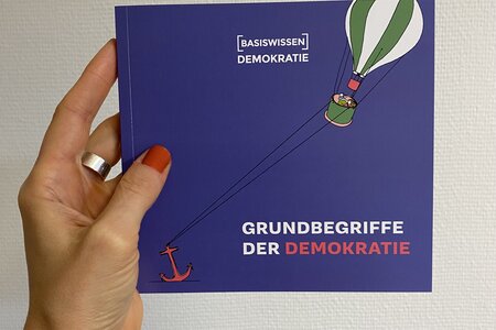 Eine Hand hält das Basiswissen Grundbegriffe der Demokratie vor eine weiße Wand. 