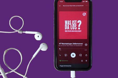 Ein Smartphone mit Kopfhörern. Das Display zeigt den Podcast "Was ist da los? Über Politik und Gesellschaft" der Landeszentrale. 