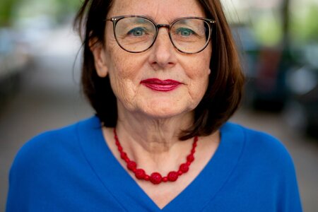 Die Frau trägt einen blauen Pullover, eine rote Kette, eine Brille und hat schulterlanges braunes Haar.