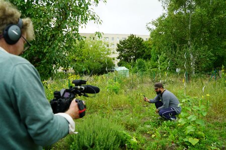 Das Bild zeigt einen Mann in seinem Garten und einen anderen Mann, der den Mann dabei mit der Kamera filmt. Im Hintergrund sind Plattenbauten einer Stadt.