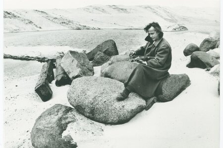Person mit Mantel sitzt auf großen Felsen in einer sandigen Landschaft