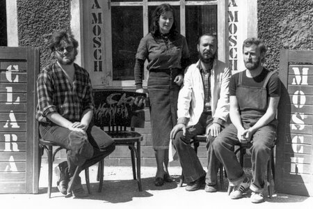 Vier Personen vor einem Gebäude - das Atelier der Gruppe "Clara Mosch" - mit den Schriftzügen 'CLARA' und 'MOSCH' auf Holztafeln.