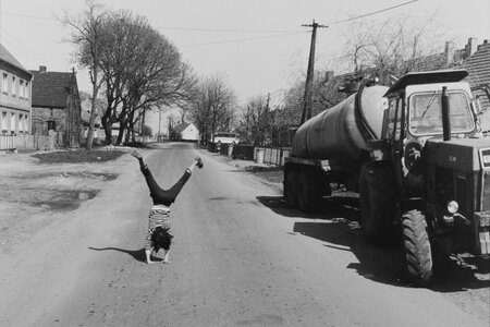 Links auf der schwarz-weiß Fotografie macht ein Kind ein Radschlag auf einer Dorfstraße. Rechts daneben steht ein Traktor am Straßenrand.