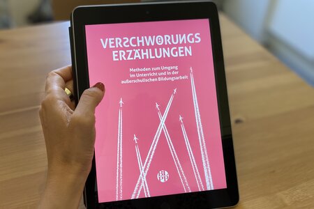 Eine Hand hält ein Tablet. Das Tablet zeigt das Bildungsmaterial "Verschwörungserzählungen. Methoden zum Umgang im Unterricht und in der außerschulischen Bildungsarbeit".