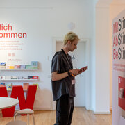 Eine Person steht in der Ausstellung "MitStimmen, AbStimmen, BeStimmen" der Landeszentrale und schaut auf Karte, die sie in der Hand hält.