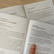 Die Verfassung des Landes Brandenburg und Grundgesetz liegen aufgeschlagen auf einem Tisch. Ein Finger zeigt auf Artikel 7a (Schutz des friedlichen Zusammenlebens) der Verfassung des Landes Brandenburg. 