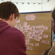 Zwei Personen stehen vor einer Pinnwand, an der viele Zettel hängen. 