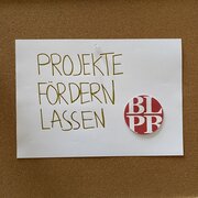 An einer Pinnwand hängt ein Zettel mit der Aufschrift: "Projekte fördern lassen". Daneben klebt das Logo der Landeszentrale.