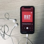 Ein Smartphone mit Kopfhörern liegt auf einem Tisch. Das Dispaly zeigt den Podcast "Was ist da los? Über Politik und Gesellschaft" der Landeszentrale. 