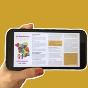 Eine Hand hält ein Smartphone. Das Display des Smartphones zeigt die Flyer "Ich kandidiere!" der Landeszentrale. 