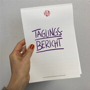 Eine Hand hält einen Notizblock hoch. Darauf steht das Wort "Tagungsbericht". 