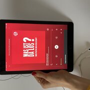 Eine Hand hält ein Tablet. Der Display der Tablets zeigt den Podcast "Was ist da los? Über Politik und Gesellschaf" der Landeszentrale.