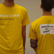 Zwei Personen in gelben T-Shirts. Auf der Vorderseite der T-Shirts steht "Wahl-O-Mat. Brandenburg" und auf der Rückseite "Willst du mit mir wählen gehen?". 