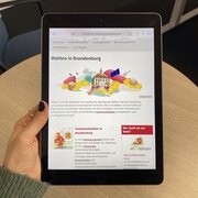 Eine Hand hält ein Tablet. Das Display des Tablet zeigt das Wahlportal der Landeszentrale.