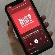 Eine Hand hält ein Smartphone. Das Display zeigt den Podcast "Was ist da los? Über Politik und Gesellschaft" der Landeszentrale.