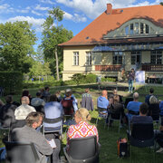 Im Hintergund die Landeszentrale. Davor steht Quiz-Master Sebastian Klussmann mit einem Mirkofon. Im Bildvordergrund sitzten die Teilnehmden des Brandenburg-Quiz. 