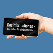 Eine Hand hält ein Smartphne. Darauf steht: "Desinformationen - eine Gefahr für unsere Gesellschaft".