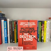 Im Hintergrund eine Reihe Bücher. Im Vordergrund das Buch „Putins Angriff auf Deutschland: Desinformation, Propaganda, Cyberattacken“ von Arndt Freytag von Loringhoven und Leon Erlenhorst.