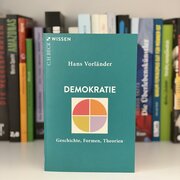 Im Hintergrund eine Reihe Bücher. Im Vordergrund das Buch "Demokratie. Geschichte, Formen, Theorien" von Hans Vorländer. 