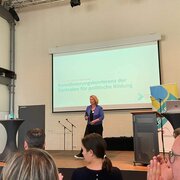 Eine Frau steht auf einer Bühne. Im Hintergrund ist eine Leinwand zu sehen. Darauf steht: "Koordinierungskonferenz der Zentralen für politische Bildung".