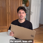 Tobias, Host der Videoreihe zu Grundbegriffen der Demokratie der Landeszentrale, sitzt am Tisch und hält ein Pappschild in der Hand.