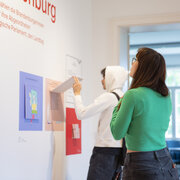 Zwei Personen stehen in der Ausstellung "MitStimmen. AbStimmen. BeStimmen." der Landeszentrale und betrachten eine Ausstellungswand.