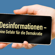 Ein Hand hält ein Handy mit der Aufschrift "Desinformationen - Gefahr für die Demokratie"