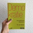 Eine Hand hält das Buch "Demokratie. Eine gefährdete Lebensform" von Till van Rahden vor eine weiße Wand.