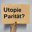 Eine Hand hält einen Zettel mit der Frage "Utopie Parität" vor eine weiße Wand.