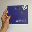 Eine Hand hält das Basiswissen Grundbegriffe der Demokratie vor eine weiße Wand. 