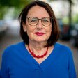 Die Frau trägt einen blauen Pullover, eine rote Kette, eine Brille und hat schulterlanges braunes Haar.