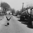 Links auf der schwarz-weiß Fotografie macht ein Kind ein Radschlag auf einer Dorfstraße. Rechts daneben steht ein Traktor am Straßenrand.